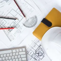 Planos de construcción, lápices, casco blanco y libreta amarilla, elementos esenciales para obtener la licencia de construcción.