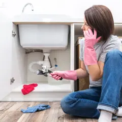 Mujer con guantes rosas y llave inglesa, revisando elementos de fontanería bajo el lavabo mientras habla por teléfono.