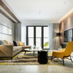 Un salón moderno con sofá marrón, alfombra llamativa y acentos amarillos. La iluminación indirecta en el techo y la pared de madera crean un ambiente acogedor, ideal para saber como iluminar un salon.