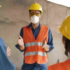 Tres trabajadores con cascos amarillos y chalecos naranjas discuten. Uno, con mascarilla, explica quién puede firmar un plan de seguridad y salud.