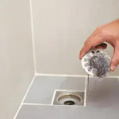 Mano retirando filtro de desagüe del baño lleno de pelos. Un paso esencial para el plano saneamiento del baño.