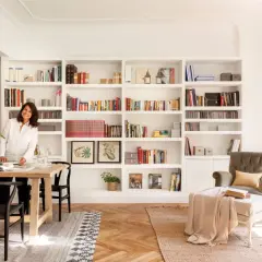 Una mujer sonríe junto a una mesa de comedor, rodeada de estanterías llenas de libros, un ejemplo de cómo los decoradores de interiores crean espacios acogedores.