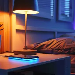 Mesita de noche con lámpara, planta y móvil cargando con luz azul. La instalación eléctrica del dormitorio crea un ambiente acogedor.