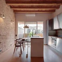 Cocina moderna con pared ladrillo visto, vigas de madera en el techo, isla central, electrodomésticos de acero inoxidable y mesa de comedor.