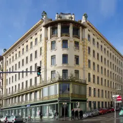 Edificio de esquina con detalles dorados, una obra de Otto Wagner. Fachada cream con ventanas oscuras y balcones verdes.