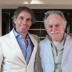 Dos hombres sonríen en un interior moderno. El de la izquierda, con traje blanco, parece un diseñador de interiores que supervisa un proyecto.
