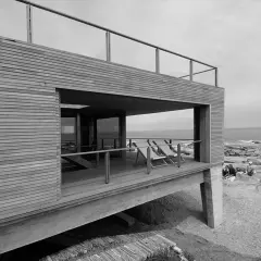 Casa de madera y hormigón con terraza y vista al mar, obra de Mathias Klotz. Personas disfrutan en las rocas.