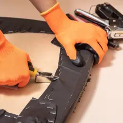 Manos con guantes naranjas usando una grapadora para tapizar una silla, fijando tela oscura a una base de madera.