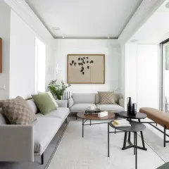 Elegante salón con sofás grises, mesas de centro negras y un banco de cuero marrón. El diseño de interiores y exteriores se fusiona con la luz natural que entra por los ventanales.