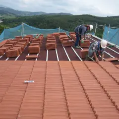 Dos trabajadores instalan tejas en la estructura de tejado a un agua. Pilas de tejas rojas esperan su colocación.
