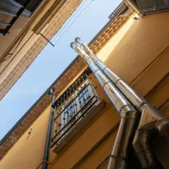 Dos tubos de acero inoxidable suben por la fachada amarilla de un edificio, parte del esquema bajantes edificio. Una ventana con rejas se ve debajo.