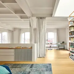 Un **espacio diafano** con vigas expuestas, estanterías de madera, zona de trabajo y cortinas blancas.