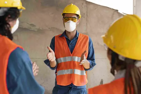 Tres trabajadores con cascos amarillos y chalecos naranjas discuten. Uno, con mascarilla, explica quién puede firmar un plan de seguridad y salud.
