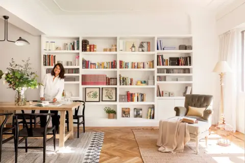 Una mujer sonríe junto a una mesa de comedor, rodeada de estanterías llenas de libros, un ejemplo de cómo los decoradores de interiores crean espacios acogedores.