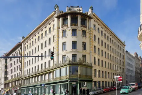Edificio de esquina con detalles dorados, una obra de Otto Wagner. Fachada cream con ventanas oscuras y balcones verdes.