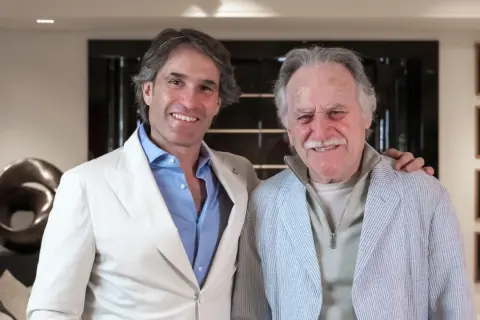 Dos hombres sonríen en un interior moderno. El de la izquierda, con traje blanco, parece un diseñador de interiores que supervisa un proyecto.