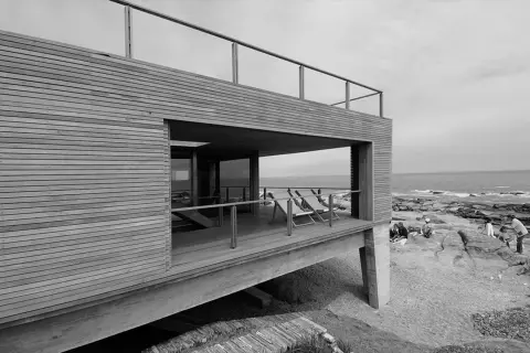 Casa de madera y hormigón con terraza y vista al mar, obra de Mathias Klotz. Personas disfrutan en las rocas.