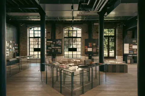 Exposición de arte y arquitectura en un espacio industrial con maquetas, planos y fotografías.