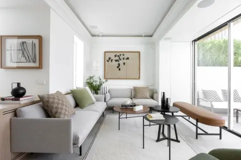 Elegante salón con sofás grises, mesas de centro negras y un banco de cuero marrón. El diseño de interiores y exteriores se fusiona con la luz natural que entra por los ventanales.