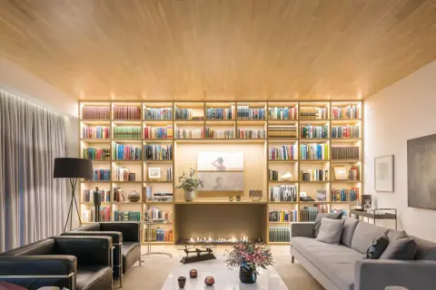 Un salón moderno con una gran librería iluminada, chimenea y sofás. Un diseñador de espacios ha creado un ambiente acogedor y elegante.