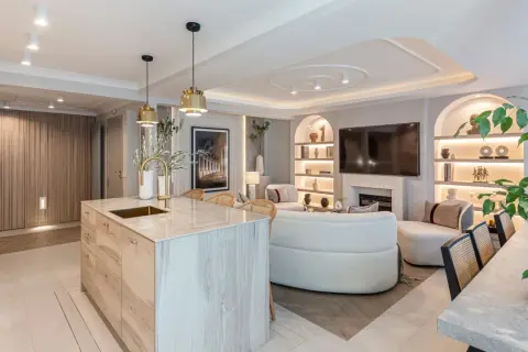 Cocina y sala de estar modernas con isla de cocina de madera, sofá curvo y estanterías iluminadas. La luz artificial crea un ambiente acogedor.