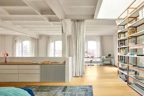 Un **espacio diafano** con vigas expuestas, estanterías de madera, zona de trabajo y cortinas blancas.
