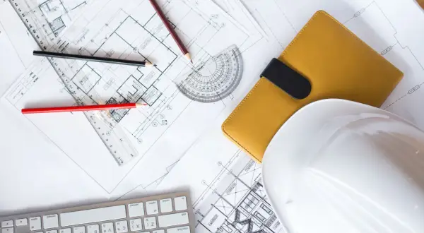 Planos de construcción, lápices, casco blanco y libreta amarilla, elementos esenciales para obtener la licencia de construcción.