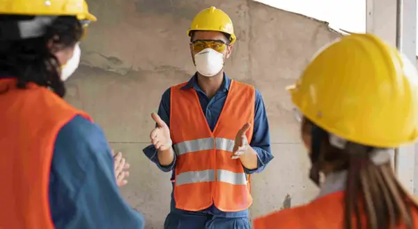 Tres trabajadores con cascos amarillos y chalecos naranjas discuten. Uno, con mascarilla, explica quién puede firmar un plan de seguridad y salud.