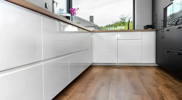 Cocina moderna con armarios blancos brillantes y encimeras de madera. El suelo vinílico SPC de madera aporta calidez y estilo.