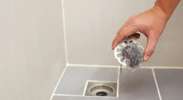 Mano retirando filtro de desagüe del baño lleno de pelos. Un paso esencial para el plano saneamiento del baño.