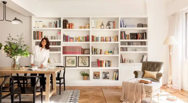 Una mujer sonríe junto a una mesa de comedor, rodeada de estanterías llenas de libros, un ejemplo de cómo los decoradores de interiores crean espacios acogedores.