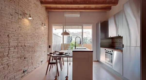 Cocina moderna con pared ladrillo visto, vigas de madera en el techo, isla central, electrodomésticos de acero inoxidable y mesa de comedor.