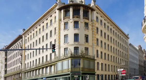 Edificio de esquina con detalles dorados, una obra de Otto Wagner. Fachada cream con ventanas oscuras y balcones verdes.