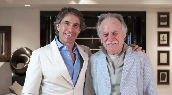 Dos hombres sonríen en un interior moderno. El de la izquierda, con traje blanco, parece un diseñador de interiores que supervisa un proyecto.