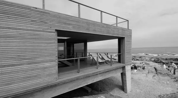 Casa de madera y hormigón con terraza y vista al mar, obra de Mathias Klotz. Personas disfrutan en las rocas.