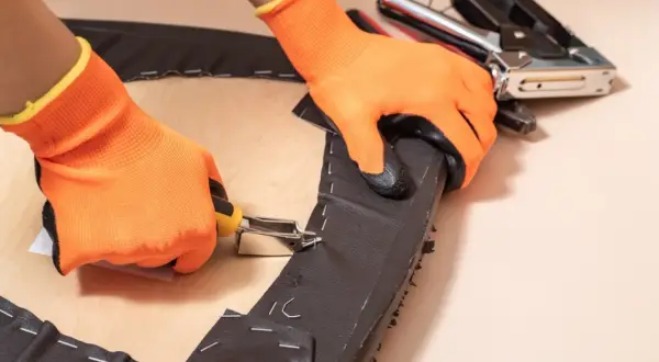 Manos con guantes naranjas usando una grapadora para tapizar una silla, fijando tela oscura a una base de madera.