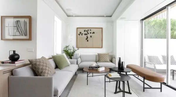 Elegante salón con sofás grises, mesas de centro negras y un banco de cuero marrón. El diseño de interiores y exteriores se fusiona con la luz natural que entra por los ventanales.
