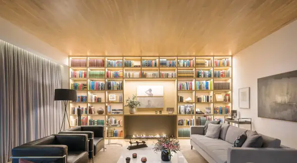 Un salón moderno con una gran librería iluminada, chimenea y sofás. Un diseñador de espacios ha creado un ambiente acogedor y elegante.