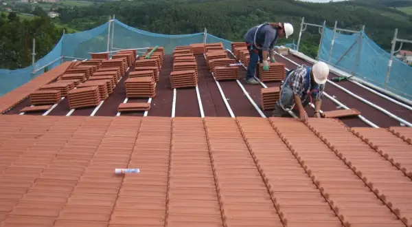 Dos trabajadores instalan tejas en la estructura de tejado a un agua. Pilas de tejas rojas esperan su colocación.