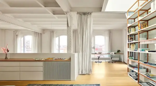 Un **espacio diafano** con vigas expuestas, estanterías de madera, zona de trabajo y cortinas blancas.