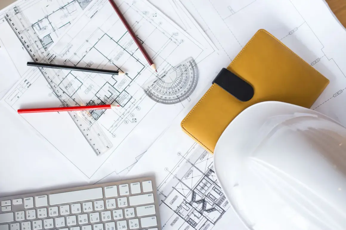 Planos de construcción, lápices, casco blanco y libreta amarilla, elementos esenciales para obtener la licencia de construcción.