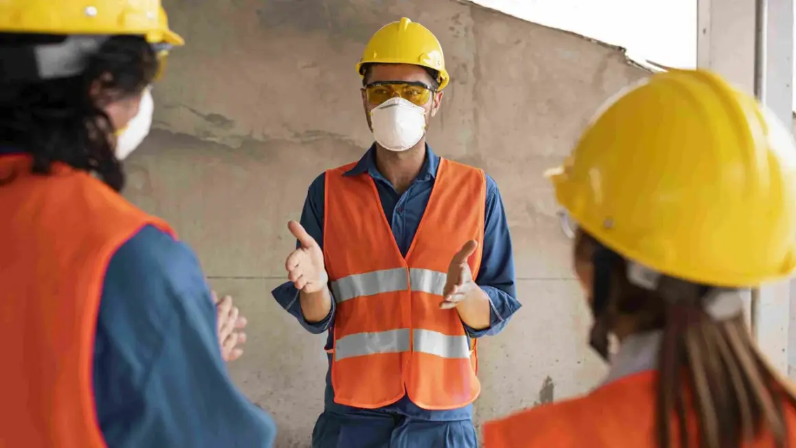 Tres trabajadores con cascos amarillos y chalecos naranjas discuten. Uno, con mascarilla, explica quién puede firmar un plan de seguridad y salud.