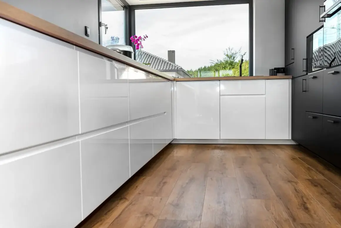 Cocina moderna con armarios blancos brillantes y encimeras de madera. El suelo vinílico SPC de madera aporta calidez y estilo.