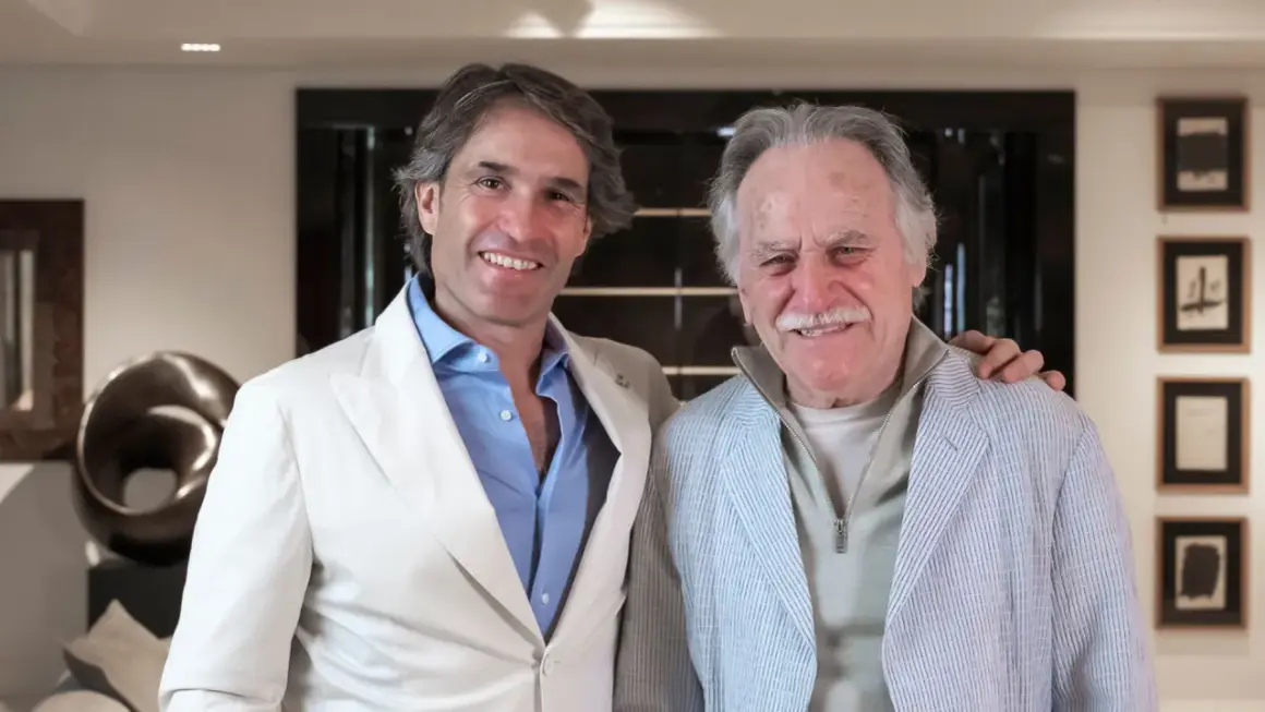 Dos hombres sonríen en un interior moderno. El de la izquierda, con traje blanco, parece un diseñador de interiores que supervisa un proyecto.