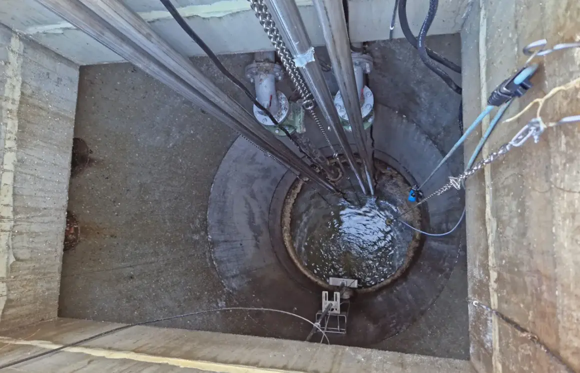 Vista superior de una estación de bombeo con tuberías y cadenas. El agua turbulenta llena el pozo, parte de la red de evacuación de aguas residuales y pluviales.
