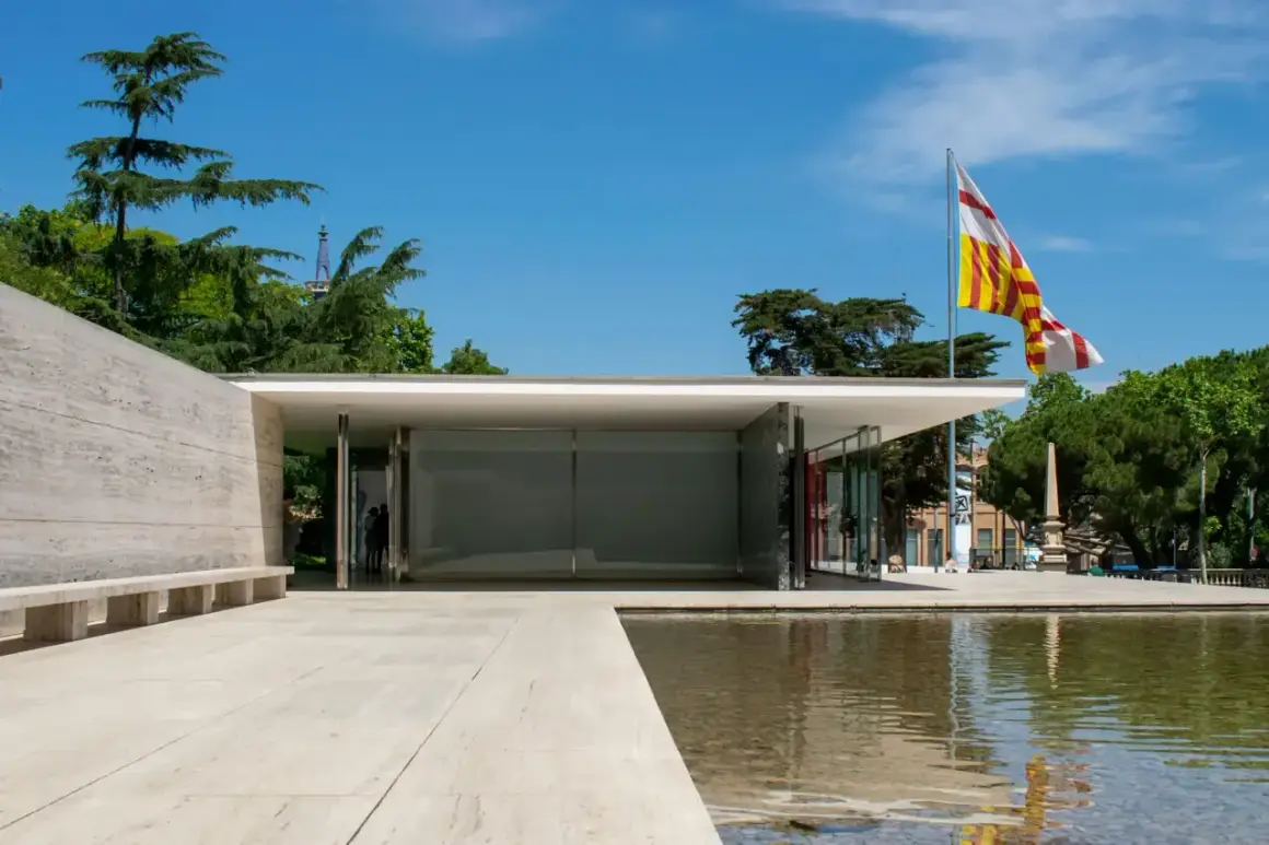 Pabellón de Mies van der Rohe, con su diseño minimalista, reflejos en el agua y una bandera catalana ondeando.