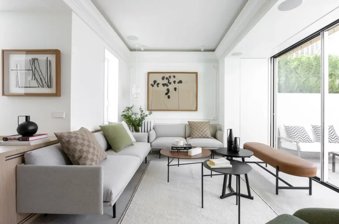 Elegante salón con sofás grises, mesas de centro negras y un banco de cuero marrón. El diseño de interiores y exteriores se fusiona con la luz natural que entra por los ventanales.