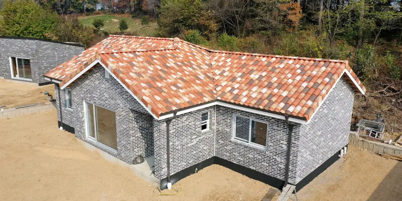 Casa con tejado de teja curva en pendiente, paredes de ladrillo gris y entorno boscoso.