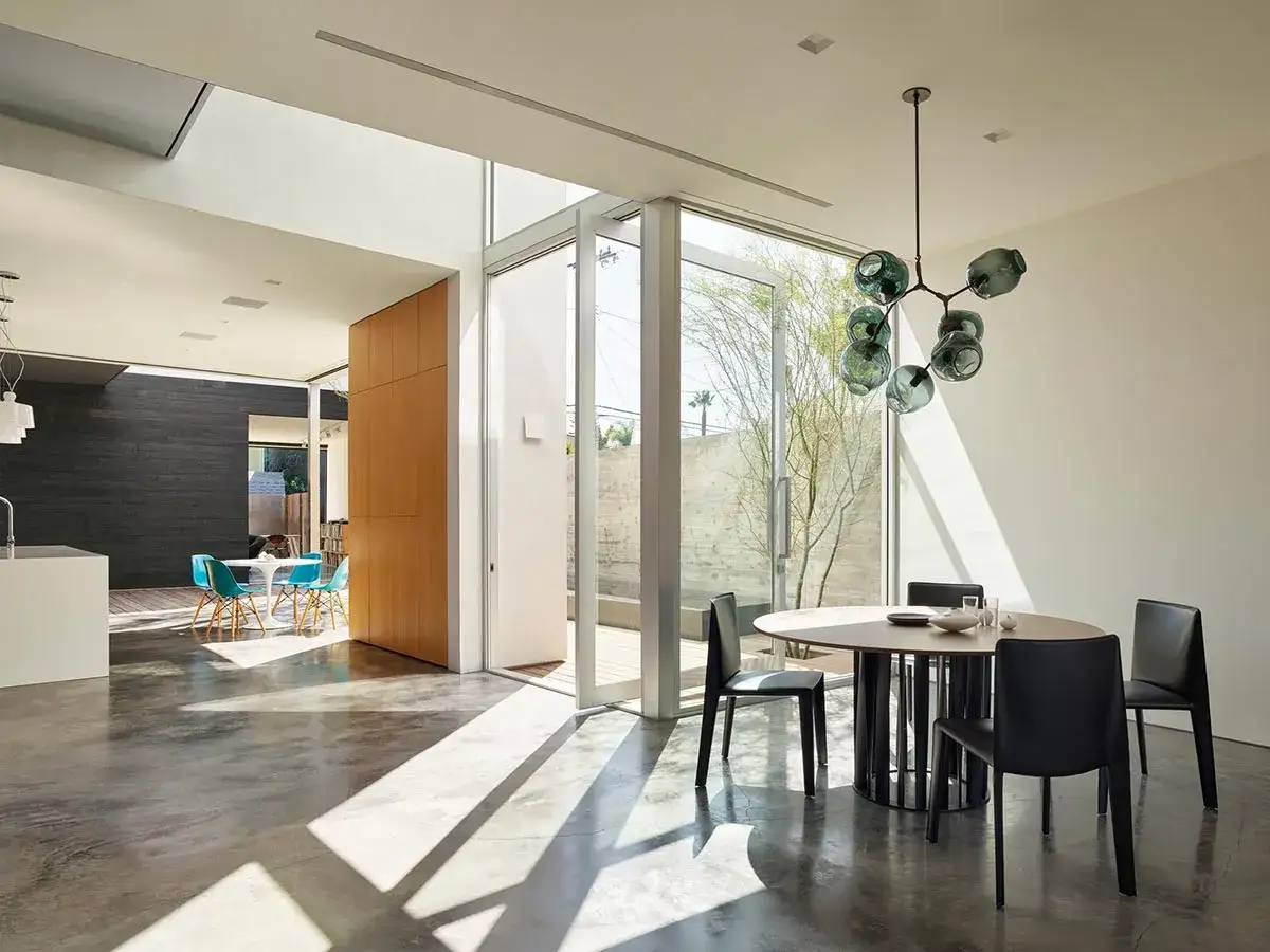 Comedor moderno con mesa ovalada y sillas negras, iluminado por varios tipos de luz natural que entran por grandes ventanales y tragaluz.