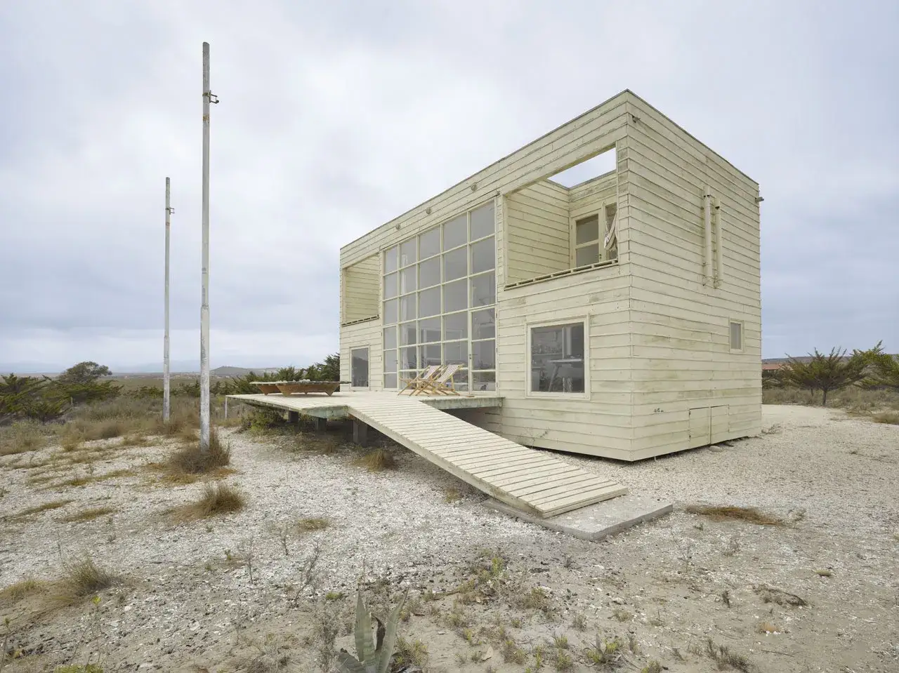 Casa de madera con grandes ventanales, dise&ntilde;ada por Mathias Klotz, en un paisaje &aacute;rido con dos postes altos.